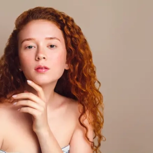 portrait of red-haired girl with long curly hair and cute freckles on beige background in the studio. Retouched photo. Skin care concept with pigmentation